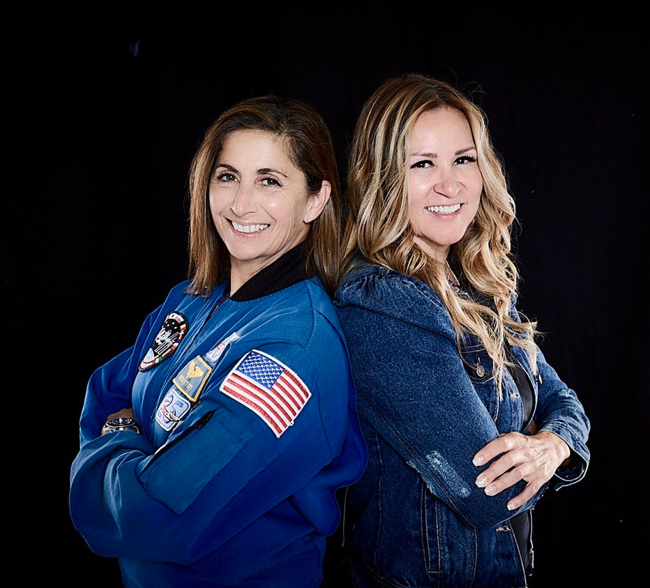 Two women, one in an astronaut suit with an American flag patch, standing back-to-back against a black background.