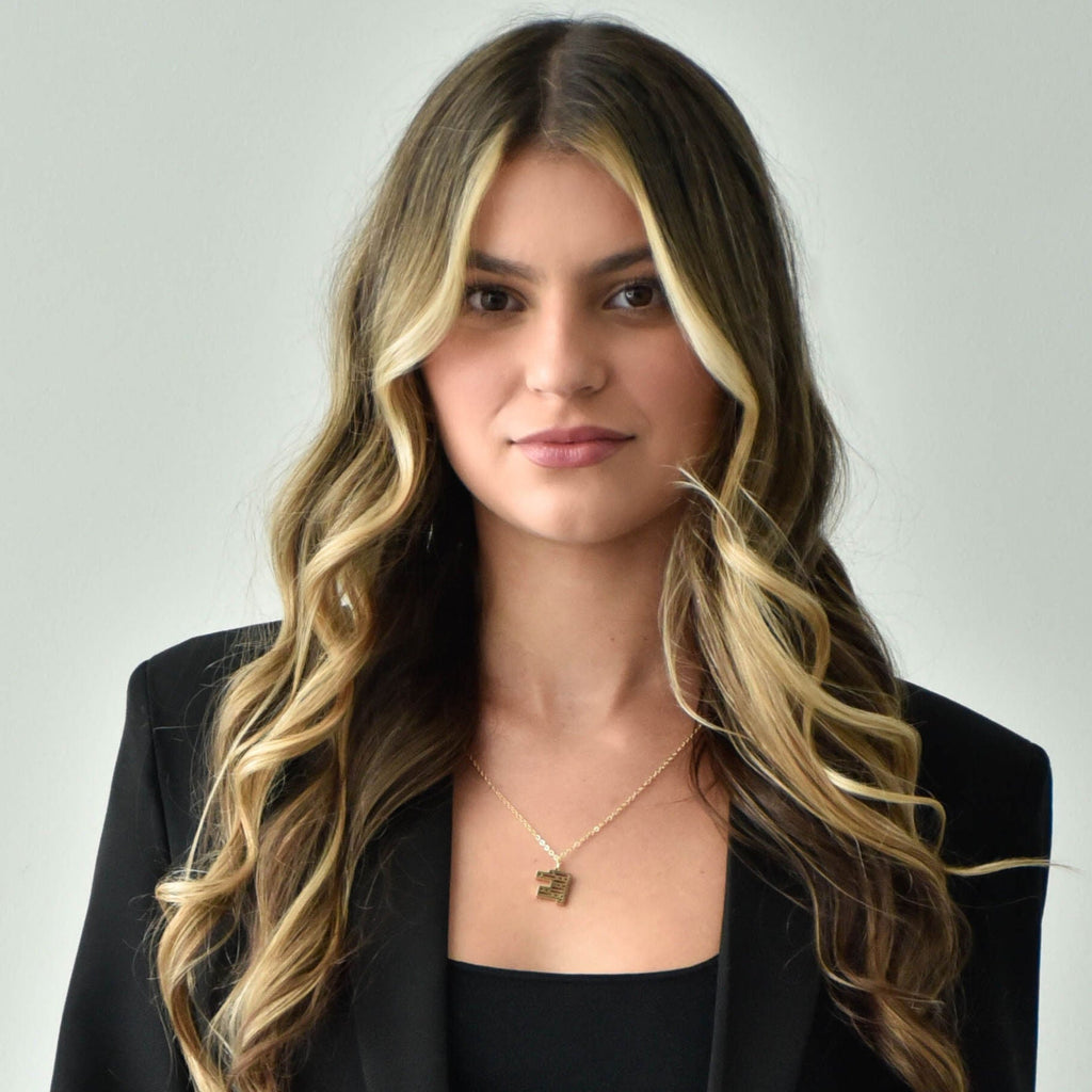 Woman wearing a gold-plated necklace in a black blazer, black top, and blue jeans against a plain background.